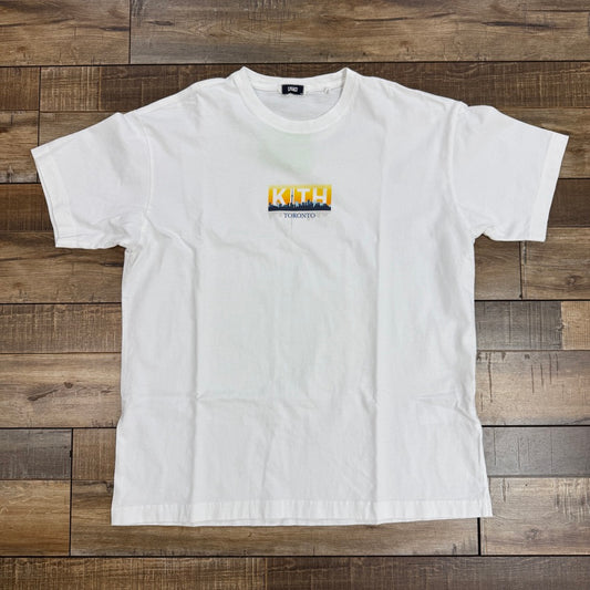 A preowned Kith Toronto Classic Logo Tee White by KITH showcases KITH with TORONTO beneath it, featuring colors inspired by the Toronto Raptors jersey. Displayed on a wooden floor, this tee captures casual yet stylish vibes for any fan.