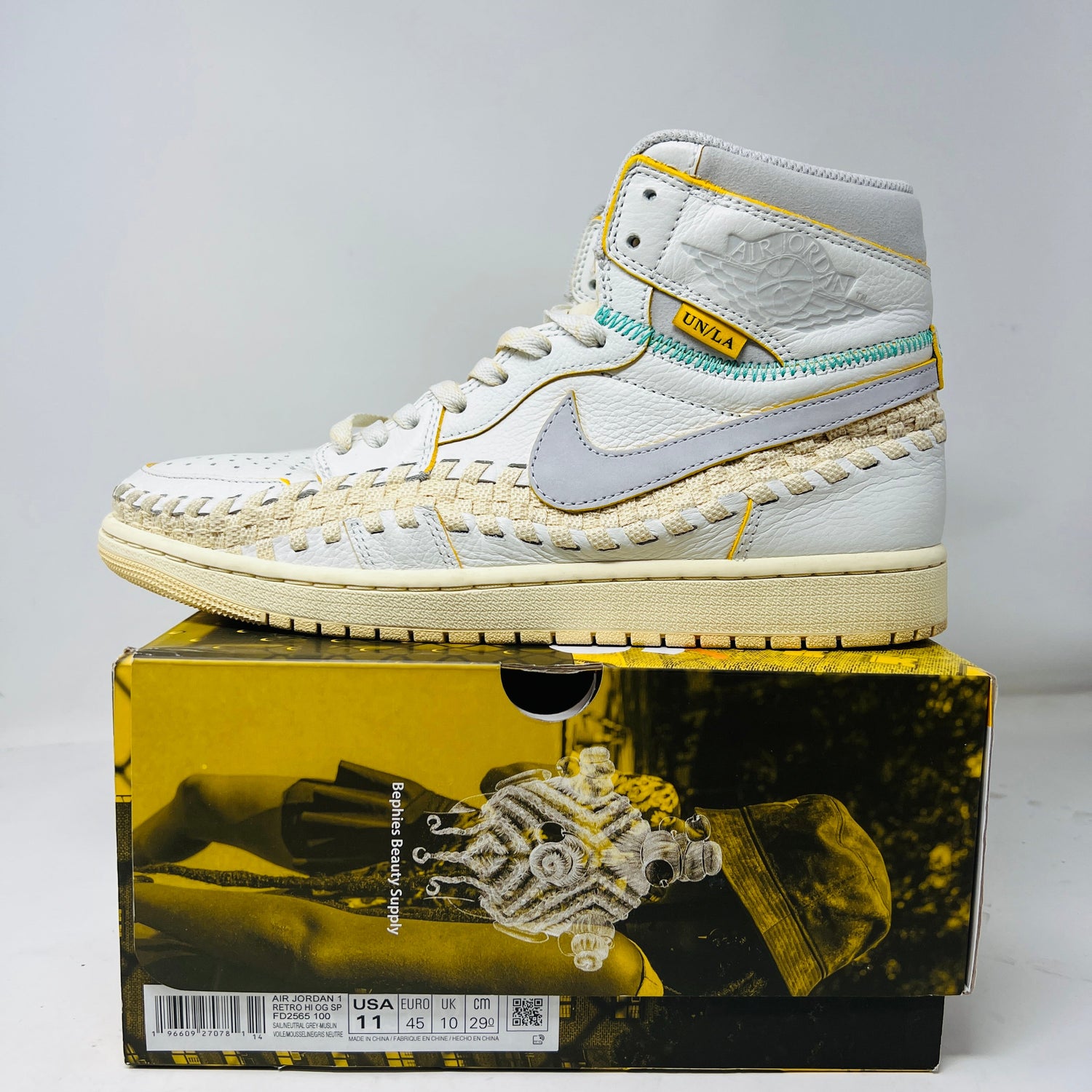 The Jordan 1 Retro High OG SP Union LA Bephies Beauty Supply Summer of ‘96 sneaker, in white and beige with a woven design and yellow accents, sits on a shoebox featuring an artistic image and UN/LA label. The Nike logo, size, and barcode are visible—ideal for light wear in 2023.