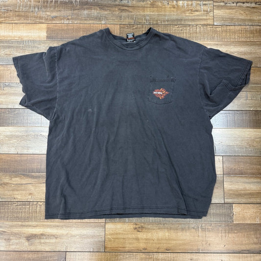 A preowned, worn Vintage Harley Davidson T-Shirt in black with short sleeves and a front pocket bearing a small faded logo is laid flat on a wooden floor, displaying slight signs of wear. .