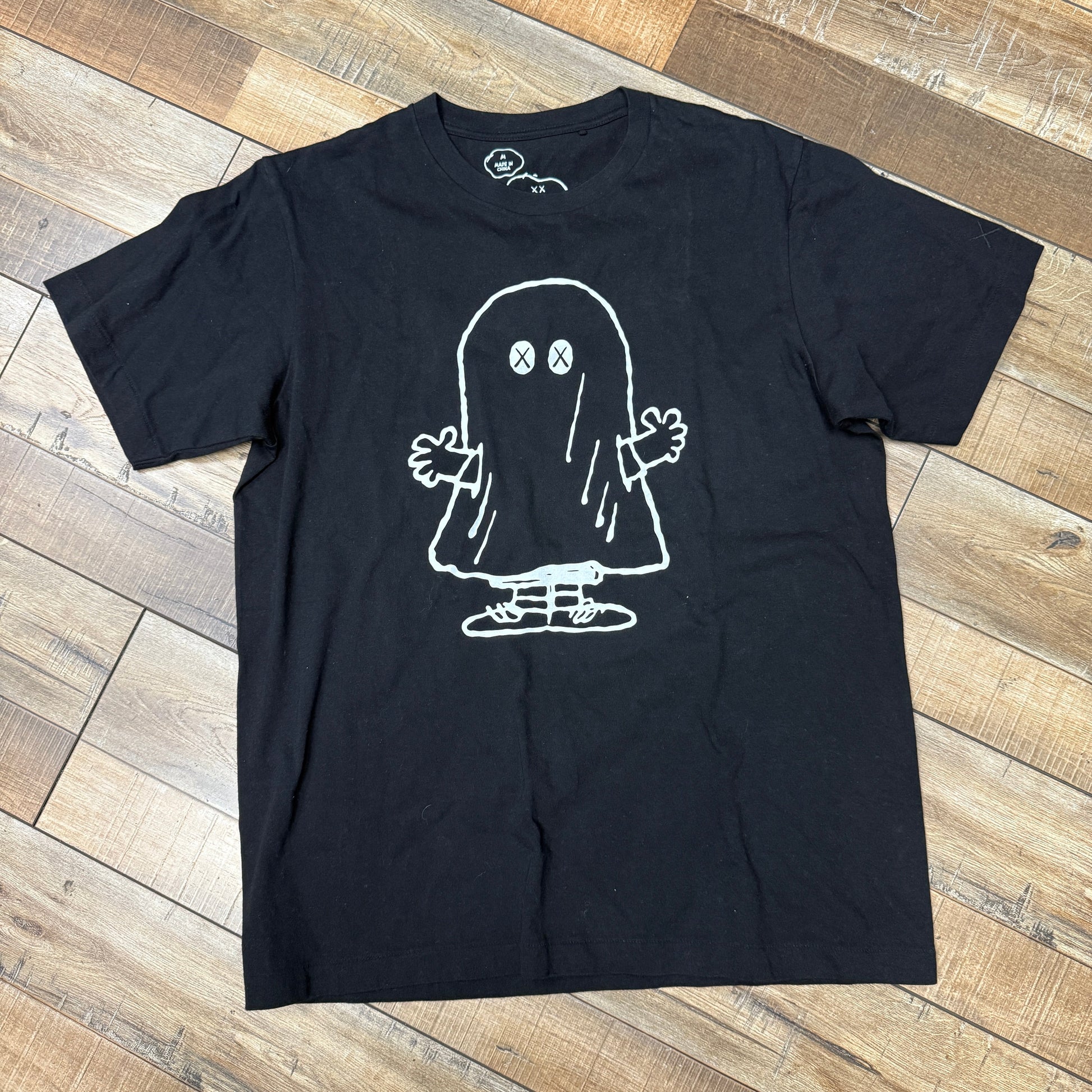 The KAWS x Uniqlo x Peanuts Ghost Tee Black, designed by Kaws, features a white outline illustration of a ghost with crossed-out eyes, gloved hands, and sneakers on a black t-shirt laid on a wooden floor.