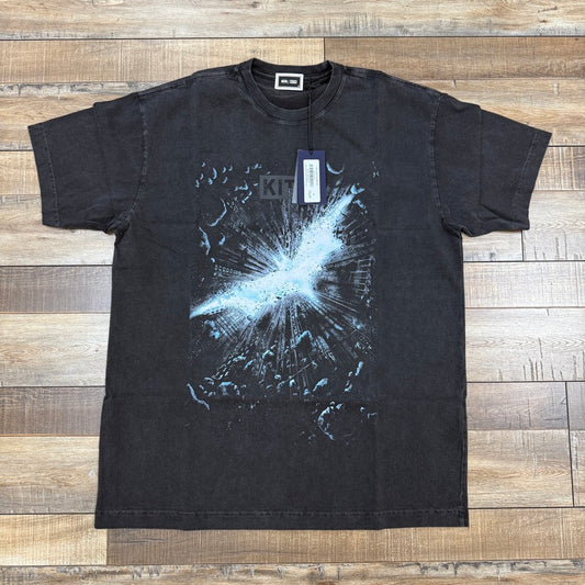 The Kith x Batman Dark Knight Adv Vintage Tee Black, a black T-shirt from KITH, features a cosmic explosion graphic in blue and white on the front. Brand new and laid flat on a wooden floor, it comes with an authenticating tag attached to the neckline.