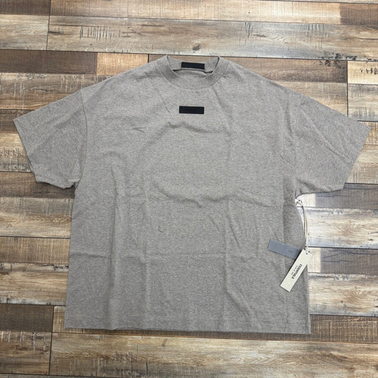 An Essentials T-Shirt Gray, featuring a black rectangular label near the neckline and a tagged sleeve, is laid flat on a rustic wooden surface that highlights its authentic design with varying tones.