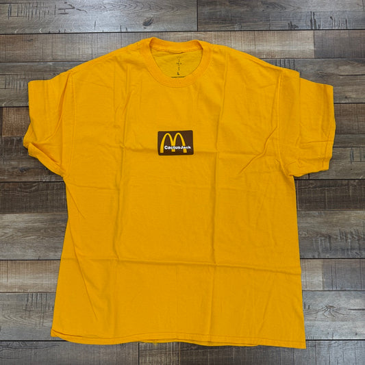 A brand new Travis Scott x McDonalds Sesame Inv II T-shirt from Cactus Jack is laid flat on the wooden floor, featuring a small, stylized logo resembling the McDonalds arches with text beneath it to showcase its authentic design.