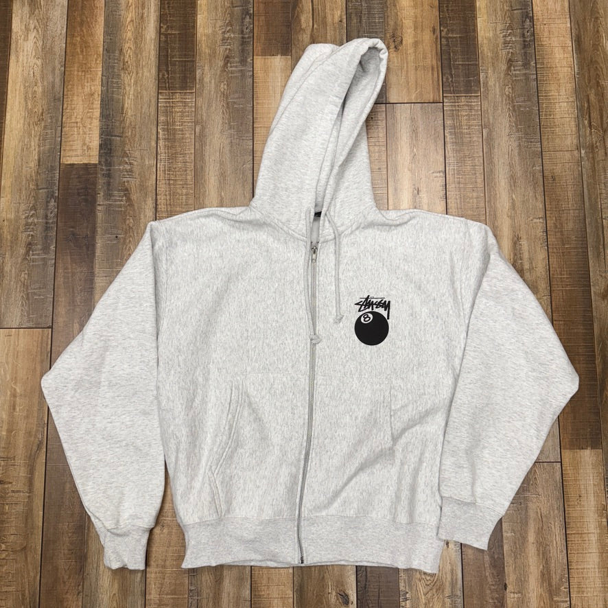 Brand new Stussy 8-Ball Zip Hoodie Grey, featuring a hood, long sleeves, and a small black emblem on the left chest. Laid flat on a wooden floor to highlight its pristine condition.