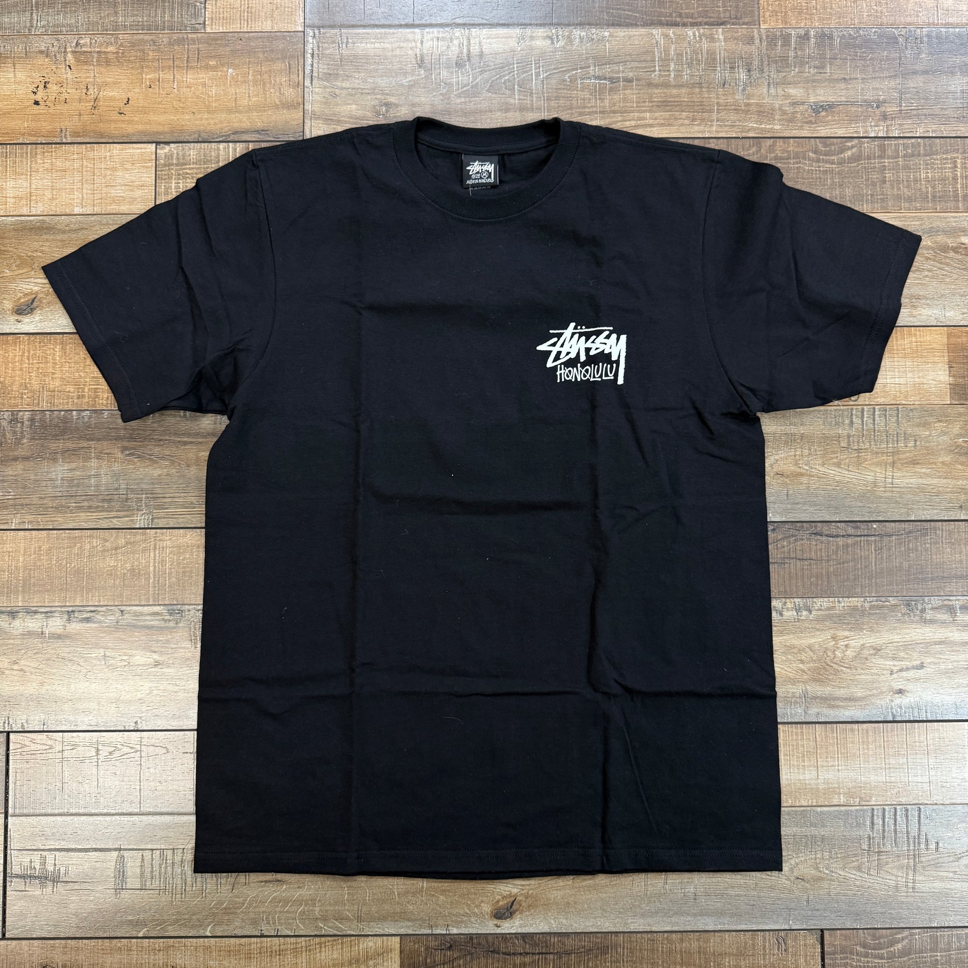 A brand new Stussy Honolulu Tee Black, featuring an authentic white Stüssy Honolulu logo on the chest, is laid flat on a wooden floor.