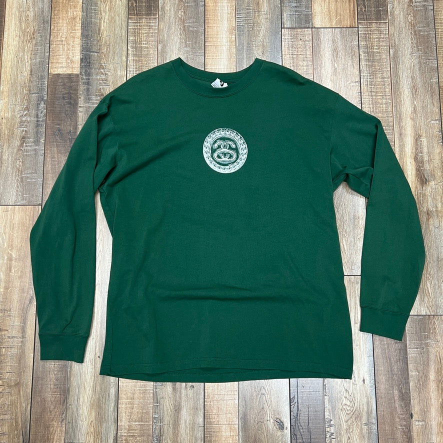 The Nike x Stussy SS Link L/S T-shirt in green, featuring a circular white logo with an abstract design and text on the chest, is laid flat on a patterned wooden floor, showcasing its pristine condition.