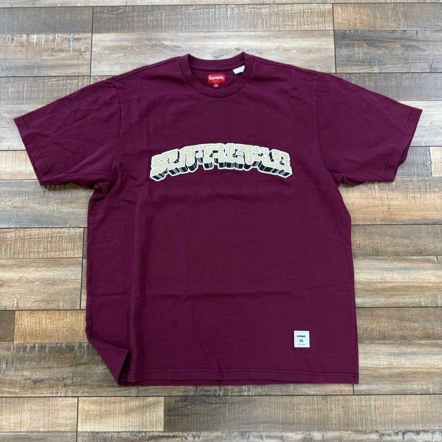 The Supreme Block Arc S/S Top in plum, a brand new t-shirt featuring bold graffiti-style text across the chest, is displayed on a wooden floor. A small square tag at the bottom right confirms its 100% authenticity in clothing/accessories.