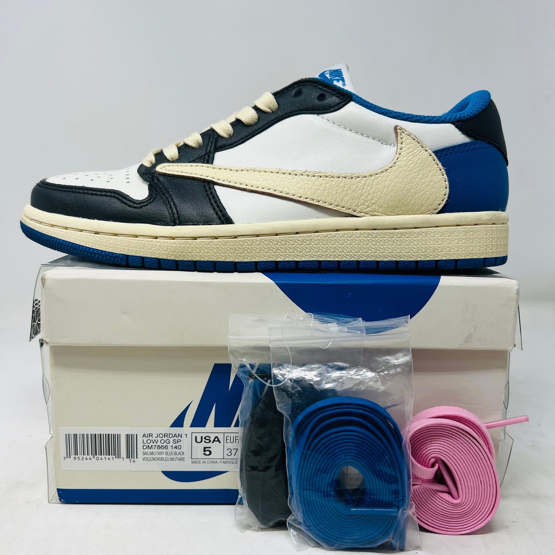 The Jordan 1 Retro Low OG SP Fragment x Travis Scott sneaker, in black, white, and blue, rests on a logo-adorned shoebox. It comes with two extra pairs of laces—one blue and one pink—neatly displayed on the white surface before the box.