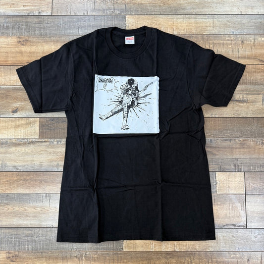 The Supreme AKIRA Yamagata Tee in black, features a white square graphic of a person kicking with impact lines and bold text. Displayed on a natural wood floor, its 100% authentic and ideal for those seeking style and originality.