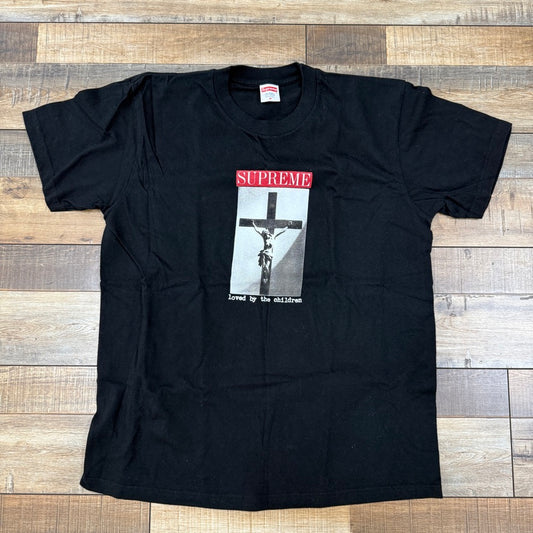 This preowned Supreme Loved By The Children Tee Black features a central crucifixion print with Supreme in bold red and loved by the children inscribed below in white. Its part of our final sales collection and is displayed on a wooden floor.