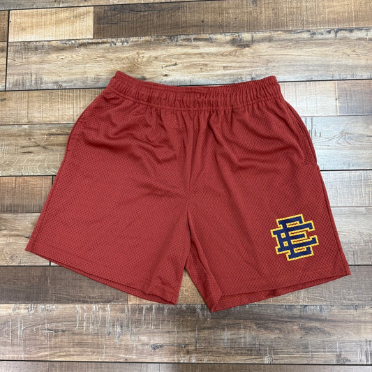 A brand new pair of Eric Emanuel EE Basic Shorts in Cinnabar, featuring a yellow and blue logo on the left leg, is lying on a wooden floor. 100% authentic.