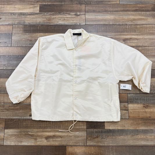 The Fear of God Essentials Coaches Jacket Cloud Dancer in cream lays flat on a wooden floor. This brand new Essentials piece has a collar, long sleeves, and a bottom hem drawstring. Its tag near the right sleeve ensures 100% authenticity.