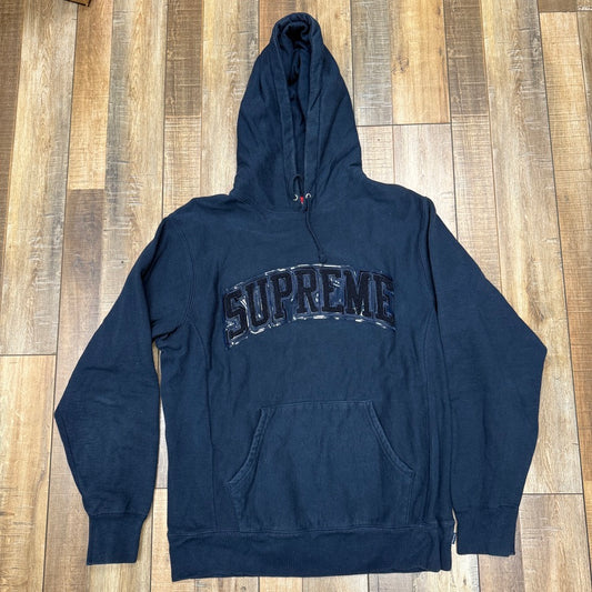 Preowned Supreme Patent/Chenille Arc Logo Hooded Sweatshirt in black with shiny SUPREME logo, shown on a wooden floor. It includes a large front pocket and drawstring hood.