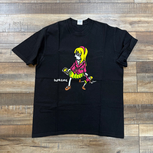 The black Supreme Suzie Switchblade Tee features a cartoon girl with blonde hair in a pink top and yellow skirt, holding a chainsaw. Supreme is displayed beside her. Laid out on a wooden floor, its an authentic edgy piece for your wardrobe.