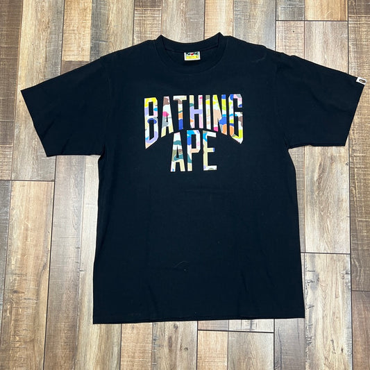 Preowned BAPE Multi Camo NYC Logo Tee in black/multicolor with reflective text, artfully displayed on a wooden floor.