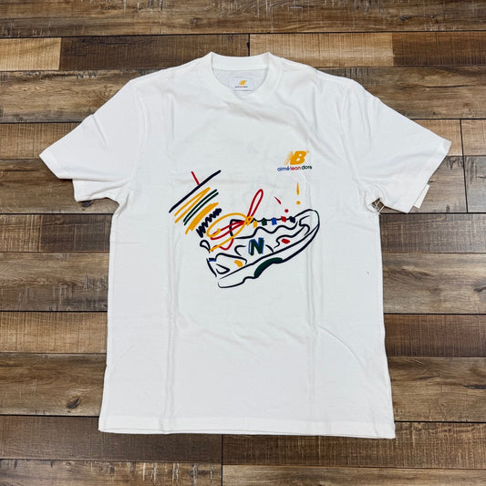 The Holy Ground Aime Leon Dore x New Balance 827 T-Shirt features a colorful abstract sneaker design in red, yellow, black, and orange on the front. Displayed on a wooden surface, this brand new product is 100% authentic with no returns or exchanges.