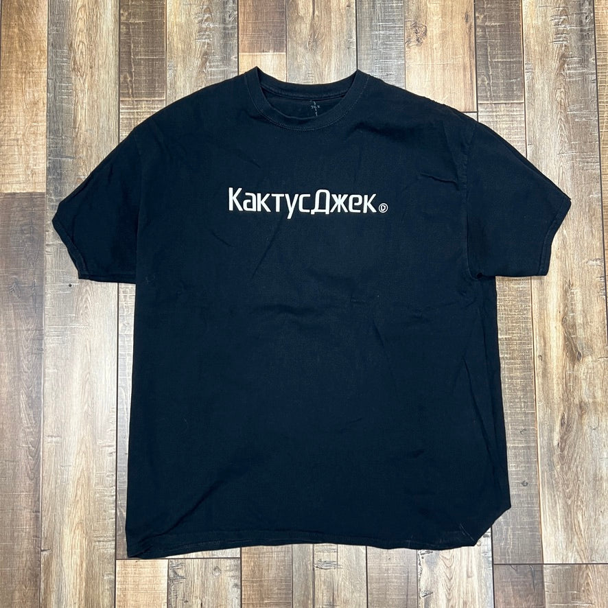 Travis Scott CJ Logo T-shirt by Cactus Jack in black, featuring white Cyrillic text on the front, laid flat on a wooden floor.