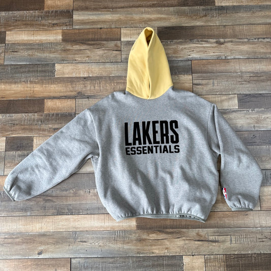 The brand new Fear of God Essentials NBA Lakers Hoodie Warm Heather, in gray with Lakers Essentials printed in black on the back and a yellow hood, is spread out on a wooden floor. This 100% authentic Essentials piece is perfect for any fan.