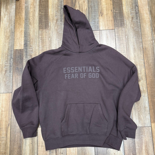 A purple raglan hoodie from Essentials, named Fear Of God Essentials, featuring ESSENTIALS FEAR OF GOD printed on the front with a front pocket, laid flat on a wooden floor.