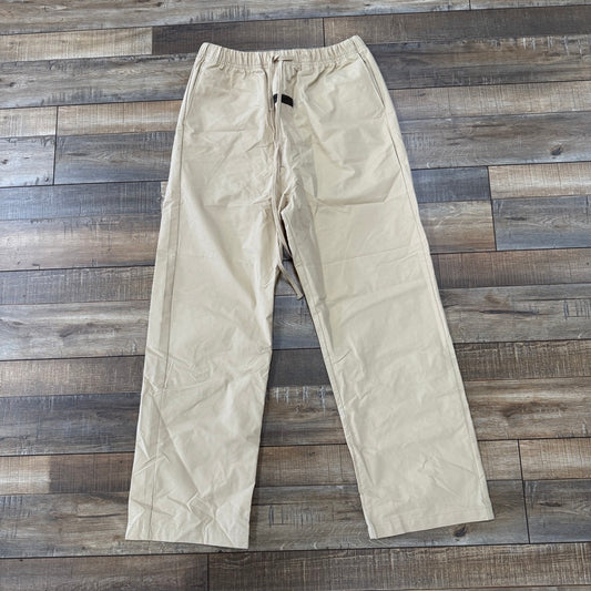 The Fear of God Essentials Relaxed Trouser in Sand, featuring an elastic waistband and drawstring, straight leg cut, and visible creases, is displayed flat on a wooden floor. It belongs to the clothing/accessories category from the Essentials brand.