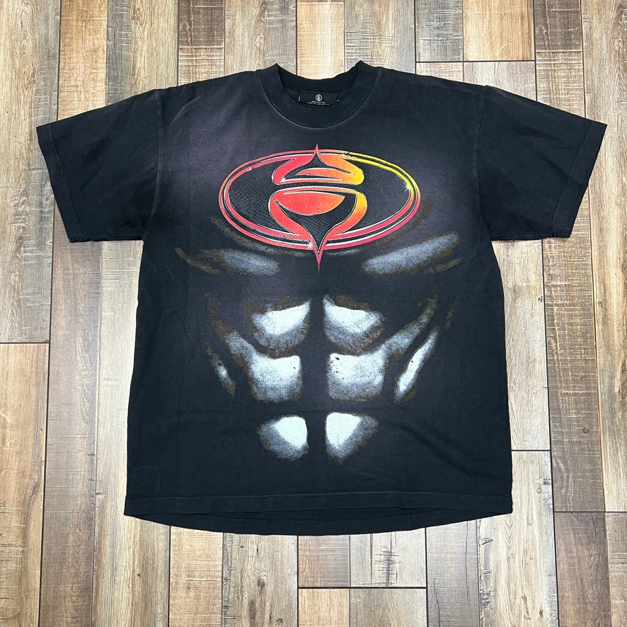 The Hellstar Superhero T-Shirt from HELL STAR is black and features an optical illusion of a muscular torso with defined abs, topped with a stylized S logo in a red-to-yellow gradient. It is displayed on a wooden floor.