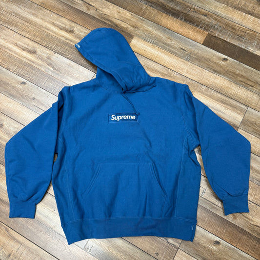 The Supreme Box Logo Hooded Sweatshirt (FW23) in blue features a white Supreme embroidery on a black chest patch, made of authentic materials. It includes a front pocket and drawstring hood, presented stylishly on a wooden floor.