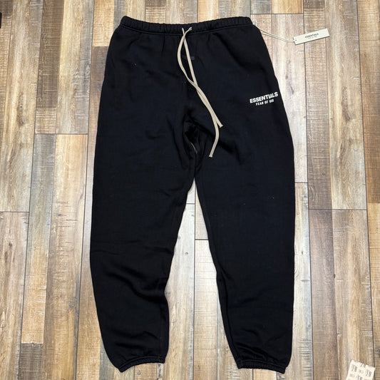 Brand new Essentials Fear of God Essentials Fleece Sweatpants in black, featuring a white Essentials Fear of God print on the left thigh. Laid flat on a wood floor, they have an elastic waistband, beige drawstring, and attached tag.