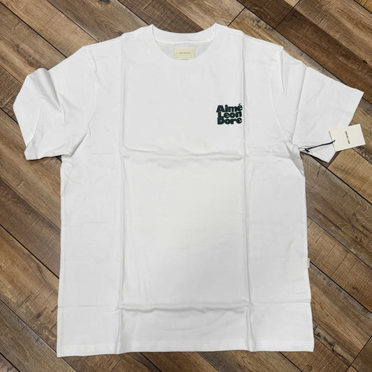 The Holy Ground Aimé Leon Dore Stacked Logo Tee in Bright White features a black Aliné Eté Bore logo on the left chest. This authentic, timeless piece lies flat on a wooden floor with a tag on the right sleeve.