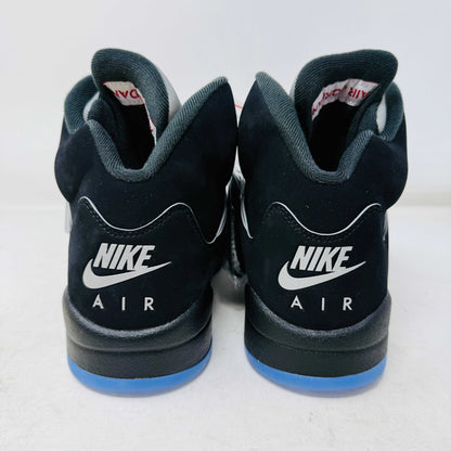 The Jordan 5 Retro OG Black Metallic Reimagined sneaker, by Jordan, comes in black with gray and silver accents, styled on a matching shoebox featuring the red 2025 Nike Air logo. This authentic shoe has a transparent sole and a signature Nike Air tag attached.