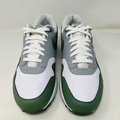 A gently used pair of Nike Air Max 1 Spiral Sage sneakers in green, gray, and white sits on a red box. Sporting the iconic swoosh and visible air sole unit, these size 12 shoes have light wear but clean uppers as noted on the label.