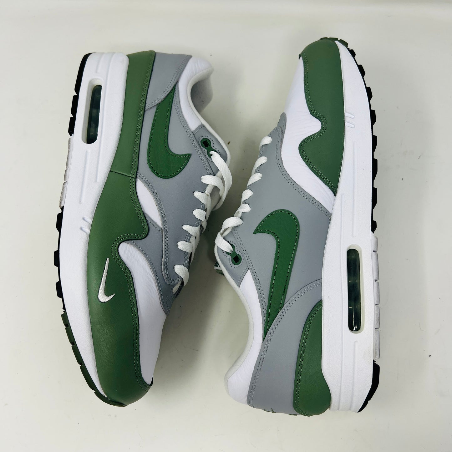 A gently used pair of Nike Air Max 1 Spiral Sage sneakers in green, gray, and white sits on a red box. Sporting the iconic swoosh and visible air sole unit, these size 12 shoes have light wear but clean uppers as noted on the label.