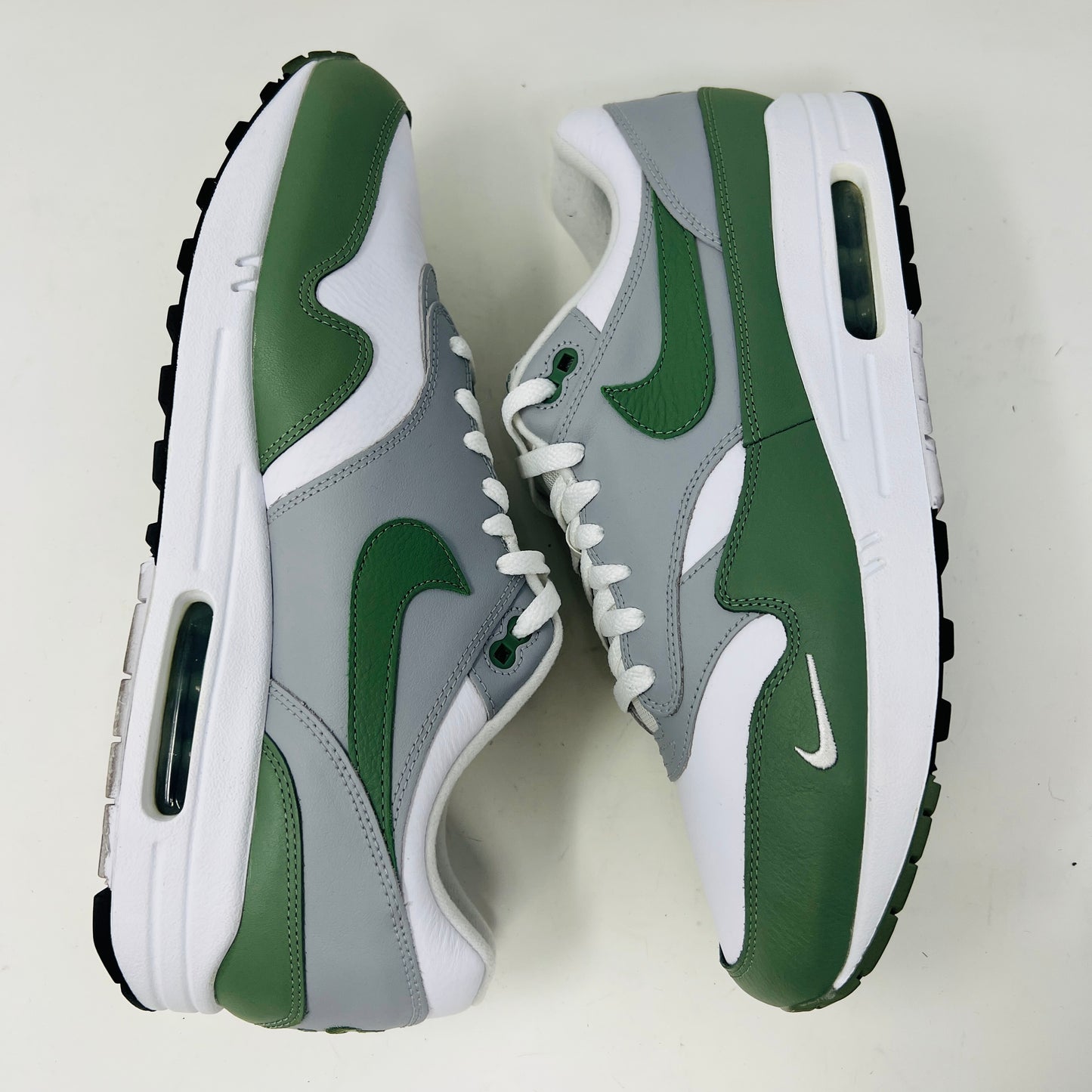 A gently used pair of Nike Air Max 1 Spiral Sage sneakers in green, gray, and white sits on a red box. Sporting the iconic swoosh and visible air sole unit, these size 12 shoes have light wear but clean uppers as noted on the label.