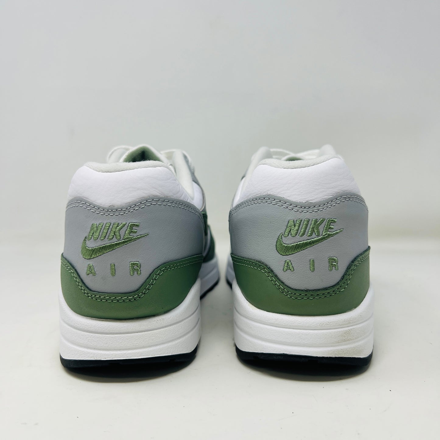 A gently used pair of Nike Air Max 1 Spiral Sage sneakers in green, gray, and white sits on a red box. Sporting the iconic swoosh and visible air sole unit, these size 12 shoes have light wear but clean uppers as noted on the label.