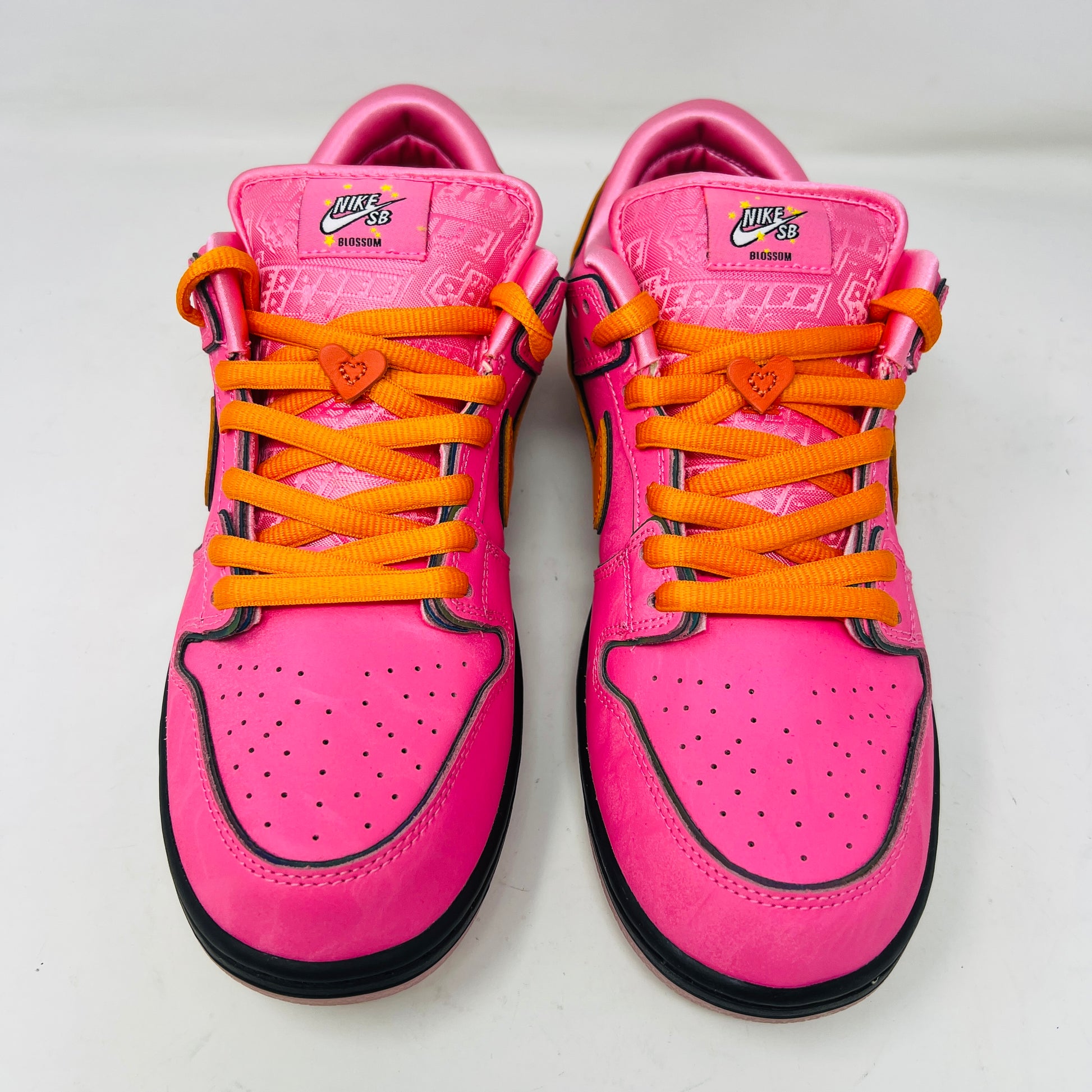 A vibrant pink Nike SB Dunk Low The Powerpuff Girls Blossom sneaker with an orange swoosh sits atop its box. It features clean uppers with black and pink details, orange laces, and comes 100% authentic, including an extra set of black laces beside the box.