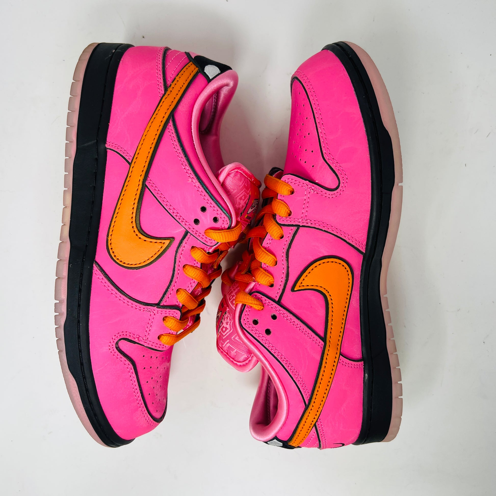 A vibrant pink Nike SB Dunk Low The Powerpuff Girls Blossom sneaker with an orange swoosh sits atop its box. It features clean uppers with black and pink details, orange laces, and comes 100% authentic, including an extra set of black laces beside the box.