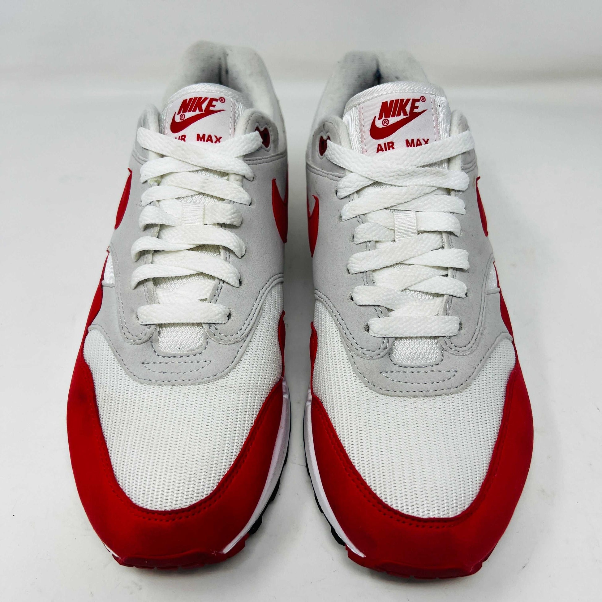 A Nike Air Max 1 Anniversary Red sneaker, with white/red uppers and a visible air unit, is displayed on an orange Nike shoebox. The box label confirms the authentic size and style details.