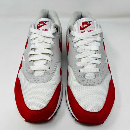 A Nike Air Max 1 Anniversary Red sneaker, with white/red uppers and a visible air unit, is displayed on an orange Nike shoebox. The box label confirms the authentic size and style details.