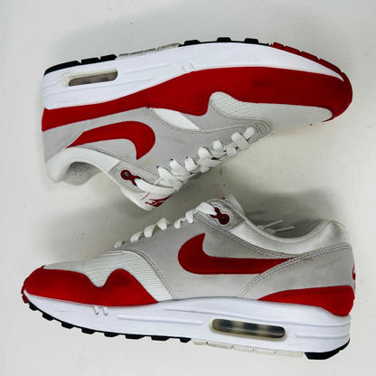 A Nike Air Max 1 Anniversary Red sneaker, with white/red uppers and a visible air unit, is displayed on an orange Nike shoebox. The box label confirms the authentic size and style details.