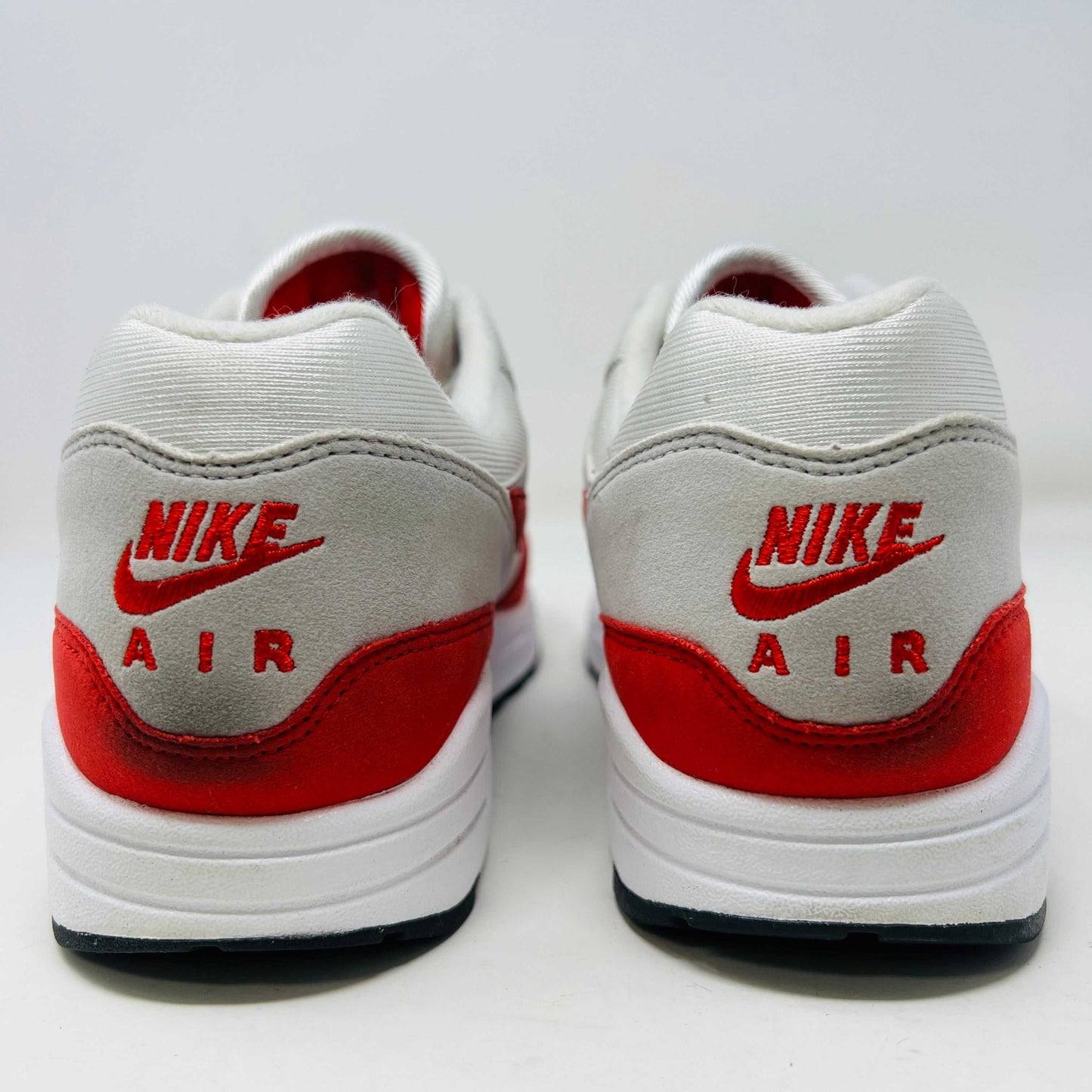 A Nike Air Max 1 Anniversary Red sneaker, with white/red uppers and a visible air unit, is displayed on an orange Nike shoebox. The box label confirms the authentic size and style details.