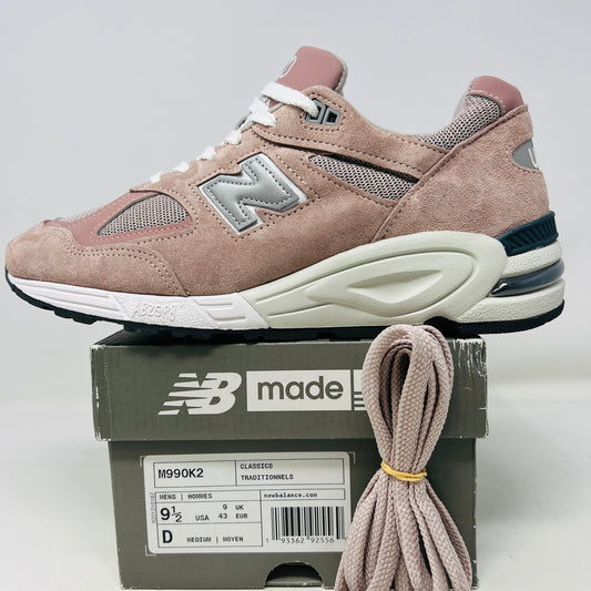 The New Balance 990v2 Kithstrike Rose sneaker from Holy Ground, in muted pink and gray, sits on its box labeled size 9.5 US, 9 UK, 43 EUR. Matching pink laces beside the box complete the authentic and stylish look with its clean uppers.