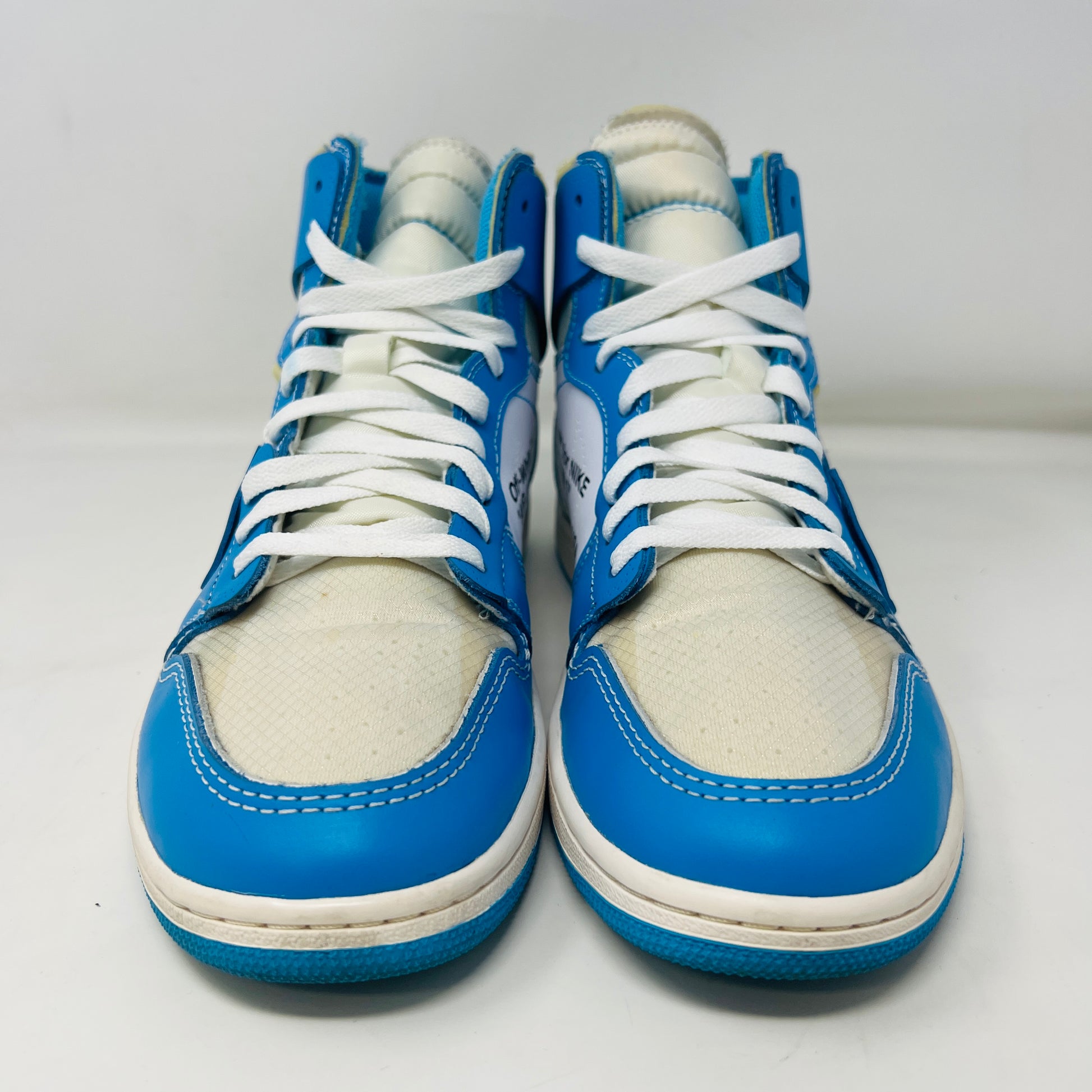 The Jordan 1 Retro High Off-White University Blue sneaker, by Jordan, in blue and white is displayed on a box with AIR text. It includes three extra laces (blue, white, orange), and a red authenticity tag against a pristine white background.