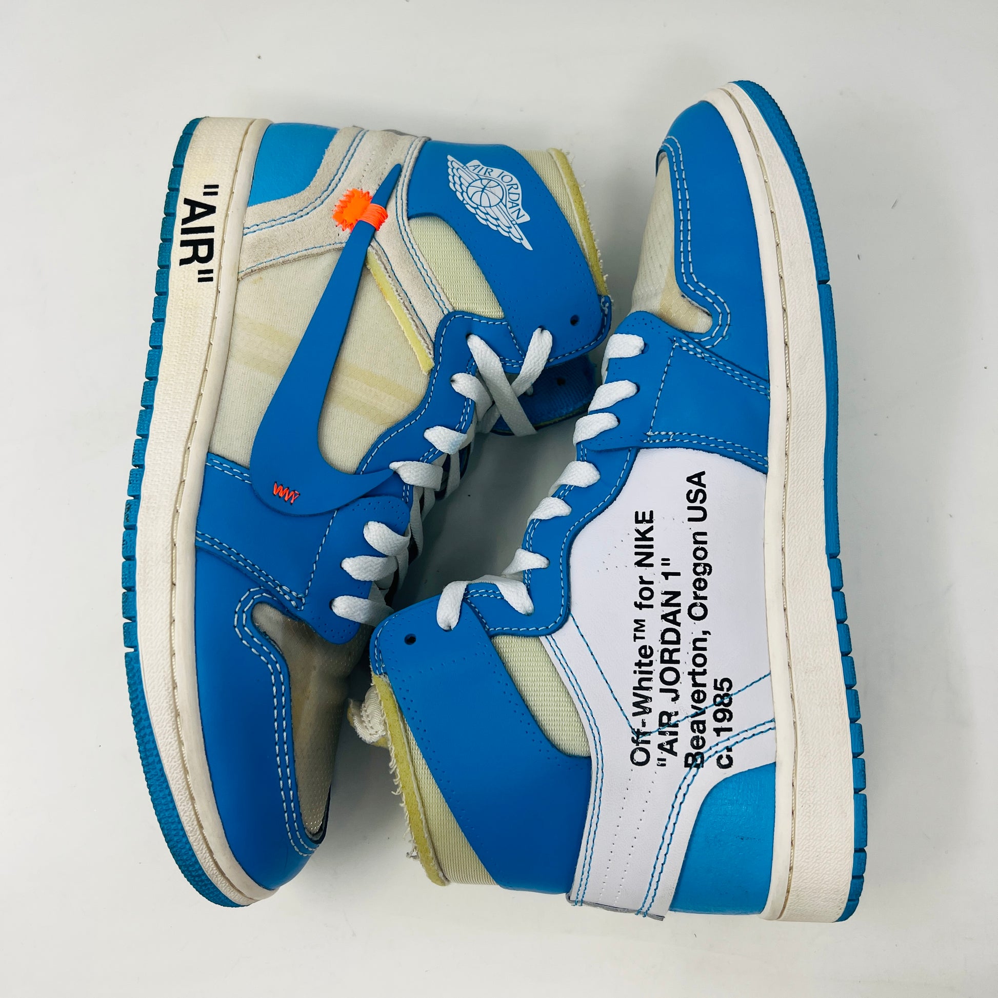 The Jordan 1 Retro High Off-White University Blue sneaker, by Jordan, in blue and white is displayed on a box with AIR text. It includes three extra laces (blue, white, orange), and a red authenticity tag against a pristine white background.