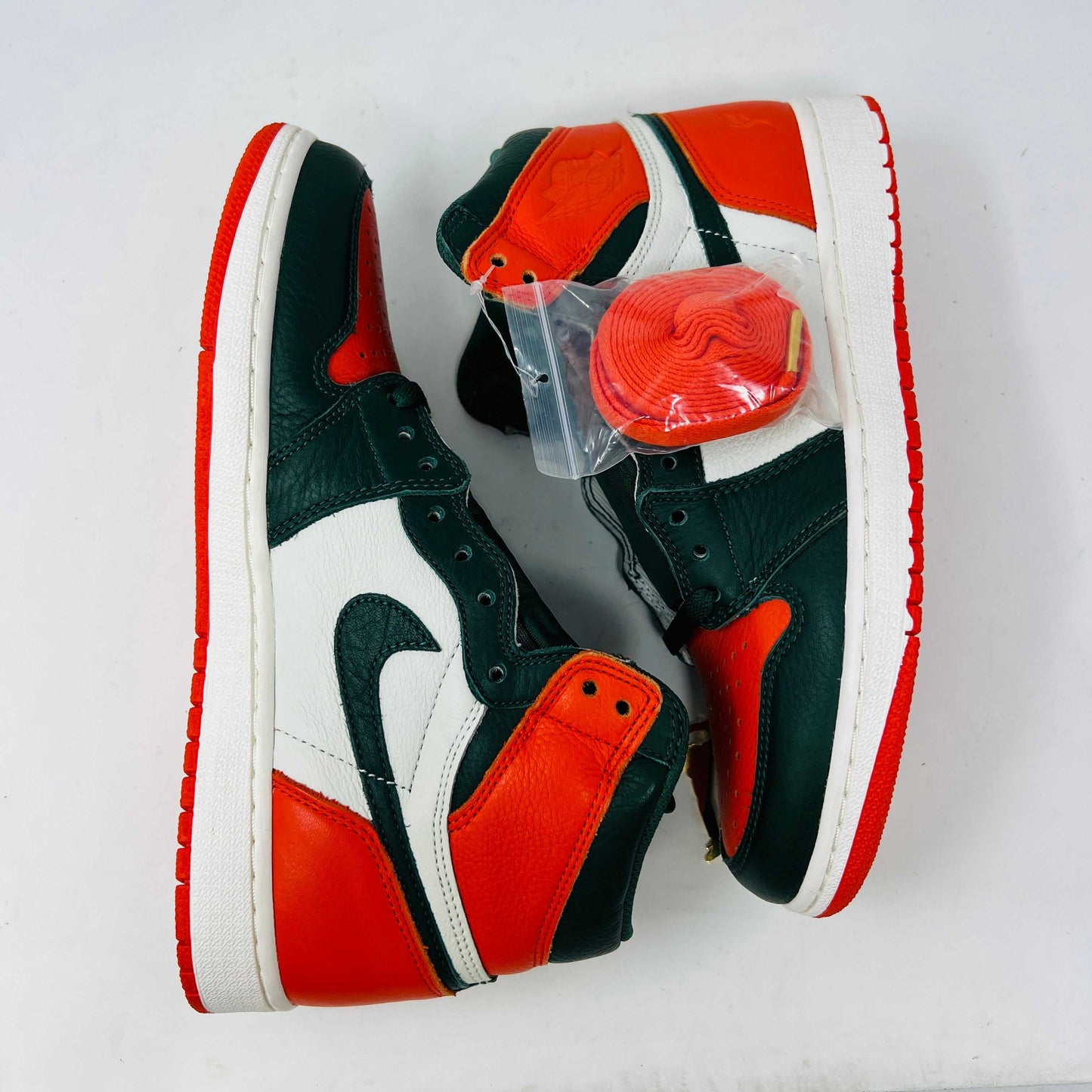 The Jordan 1 Retro High SoleFly Art Basel Sail by Jordan features orange, black, and white leather panels on a red Nike box. A small bag with bright orange laces complements the authentic sneaker, all against a plain white backdrop.