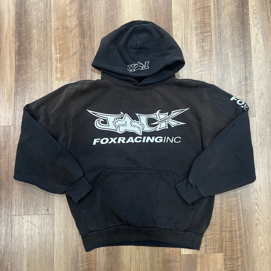 The preowned Travis Scott x Fox Racing I Hoodie Black features a front graphic of FOX RACING INC in white, W-H on the hood, and FOX on the right sleeve. Laid flat on wooden floor, it remains in excellent condition.