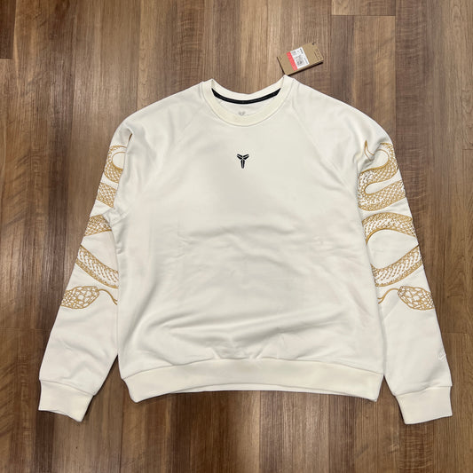 The Nike Kobe Tech Fleece Crew by KOBE is a new white sweatshirt with a small dark chest emblem and gold snake patterns on both sleeves, displayed on a wooden floor with its price tag near the neckline.