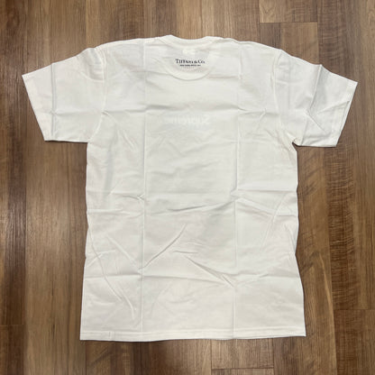 The Supreme Tiffany & Co. Box Logo Tee White, featuring a mint green logo with crisp Supreme text, is laid flat on a wooden floor in pristine condition with slight creases.