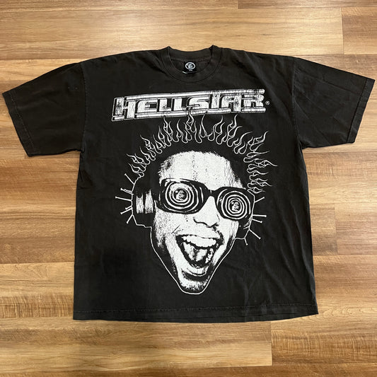 The Hellstar Rage Tee from HELL STAR is a black T-shirt with HELLSTAR in bold at the top, showcasing a graphic of a face with spiky hair, circular goggles, and an open mouth. Its stylishly laid out on a wooden floor.