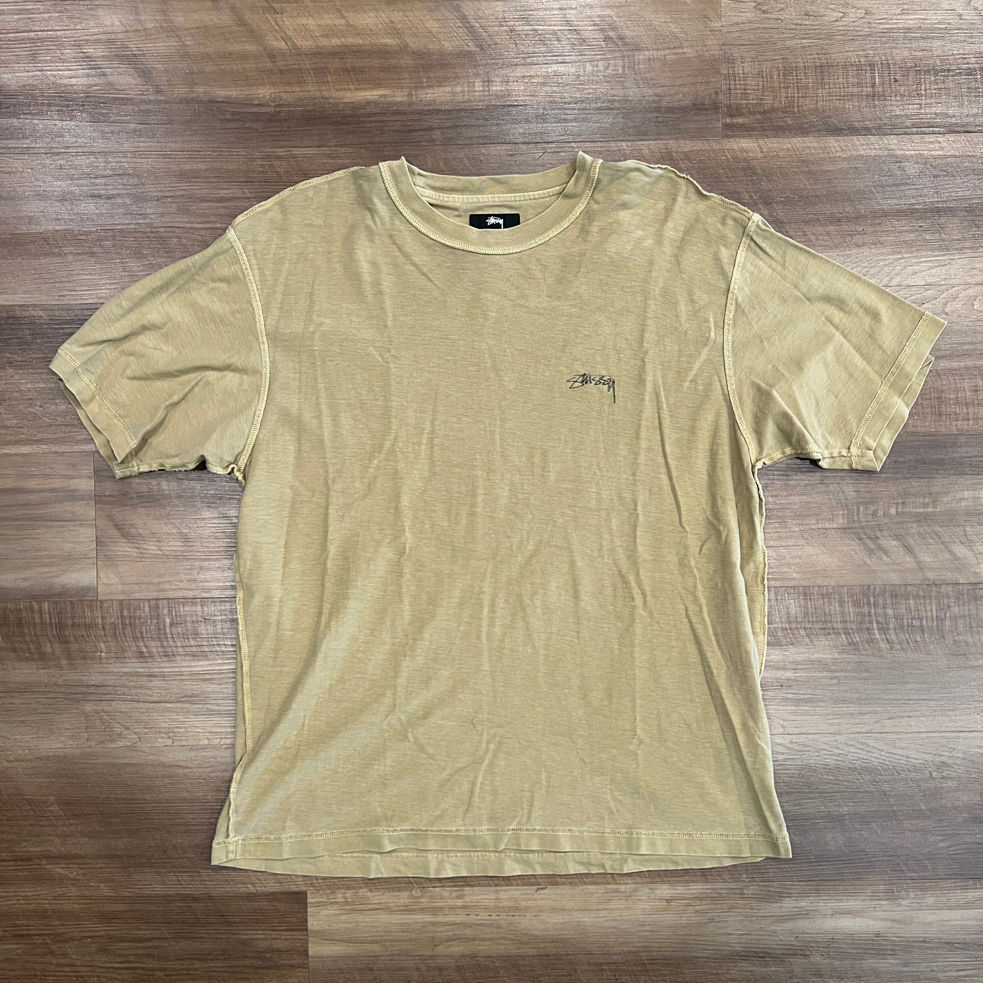 A brand new olive t-shirt from Stussy, with short sleeves and a small black logo on the left chest, is laid flat on a wooden floor.