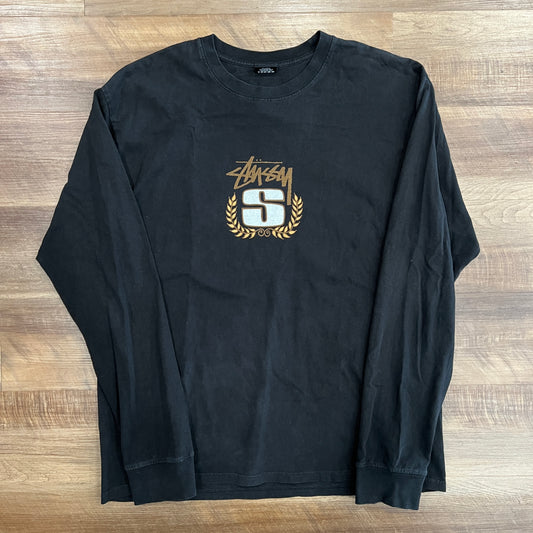 This is a preowned Stussy S Wreath Pigment Dyed Long Sleeve Black shirt with a gold and white S graphic encircled by laurel branches and a logo above, showcased flat on a wooden floor.