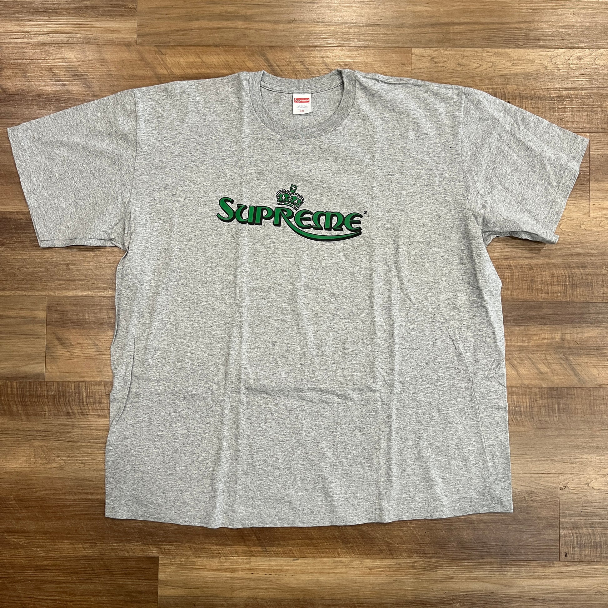 A preowned Supreme Crown Tee in Heather Grey features Supreme in green with a crown above the S, laid flat on a wooden floor.